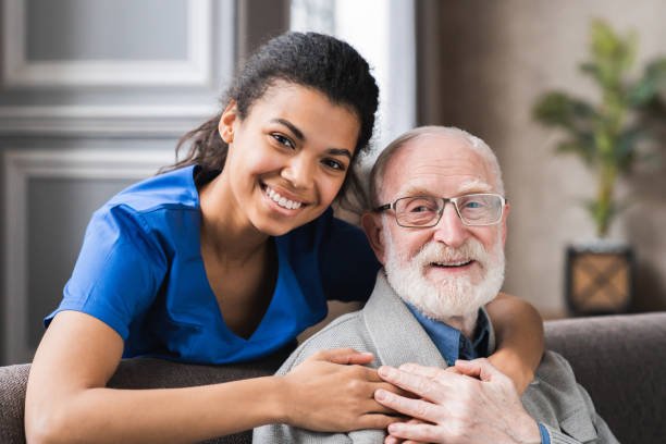 Cuidadora amigável comunicando-se com um agradável paciente do sexo masculino dos anos 80, sentados juntos no sofá. Sorridentes